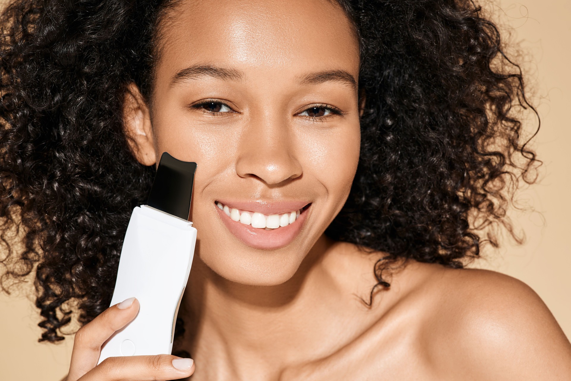 African American woman with ultrasound scrubber treating her skin for remove blackheads and cleansing facial skin, on beige background. Ultrasonic facial peeling